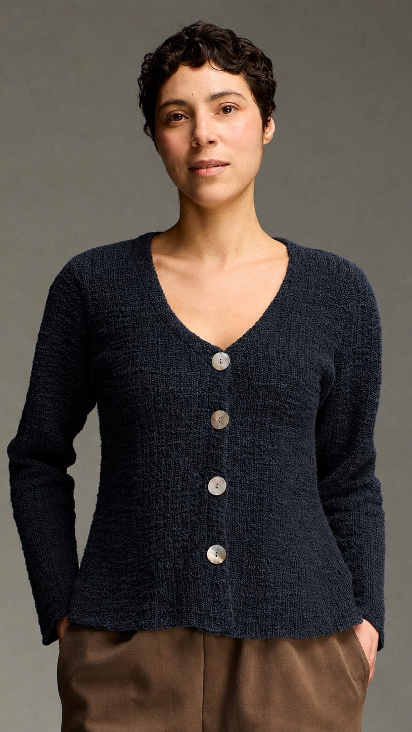 A model wearing a dark navy blue textured knit cardigan with a V-neck and silver buttons, paired with brown pants, against a solid gray studio background.