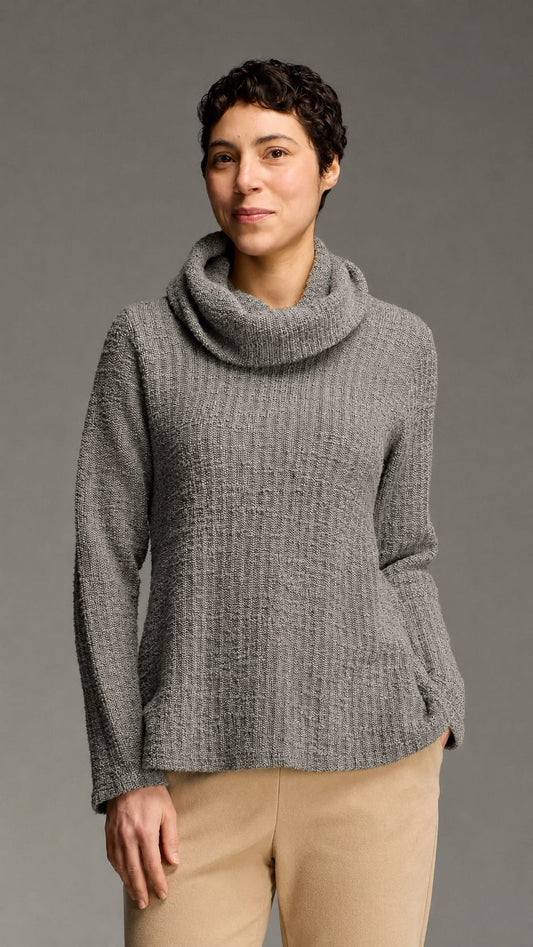 A woman wearing a textured gray cowl-neck sweater and beige pants, standing against a solid gray studio background.