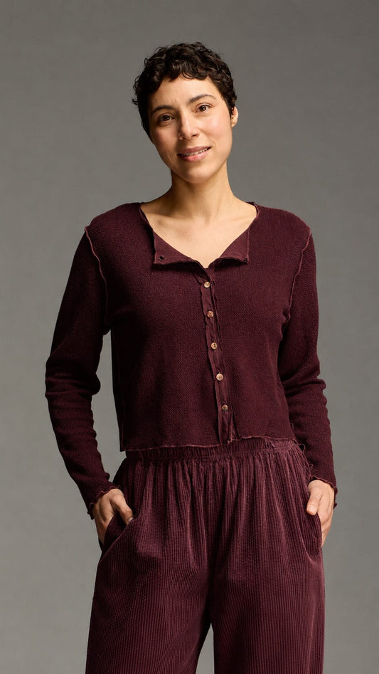 A woman modeling a matching burgundy long-sleeve button-up top and corduroy pants set against a solid gray studio background.