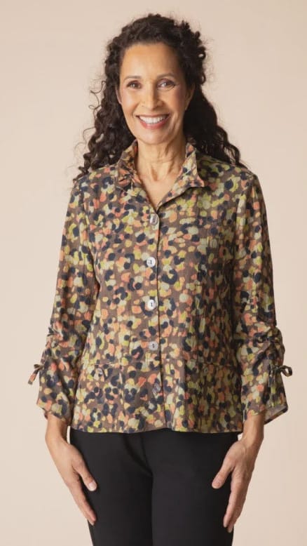 A smiling woman with curly dark hair wearing a long-sleeve button-up blouse with a multicolor abstract pattern in olive, beige, brown, and black, paired with black pants, against a solid light beige background.