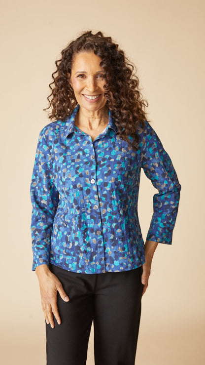 A smiling woman with curly brown hair stands against a plain beige studio background wearing a long-sleeve button-front collared blouse with an abstract blue, teal, and purple pattern, paired with black pants.
