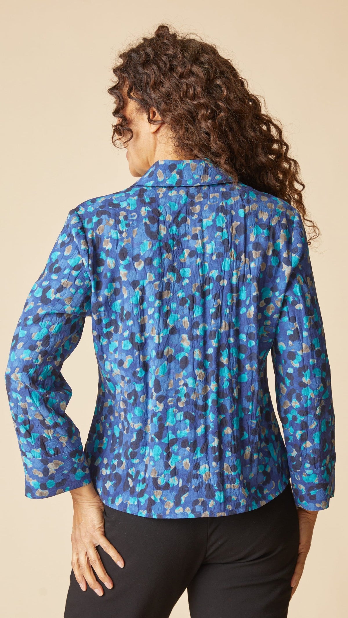 A woman shown from the back wearing a blue patterned long-sleeve blouse with black pants, set against a solid beige studio background.