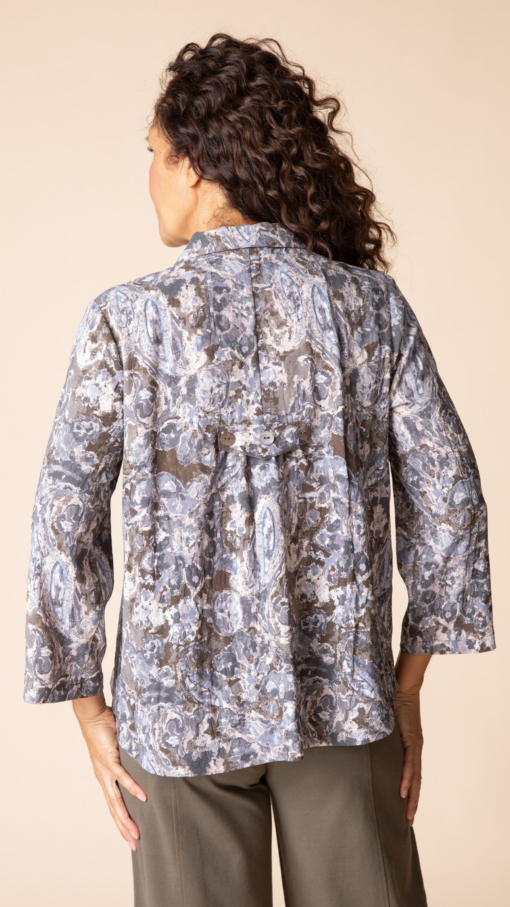 A woman shown from the back wearing a patterned button-up blouse with three-quarter sleeves and olive pants, set against a solid beige studio background.
