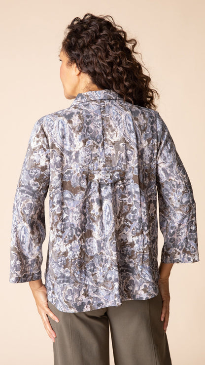 A woman shown from the back wearing a patterned button-up blouse with three-quarter sleeves and olive pants, set against a solid beige studio background.