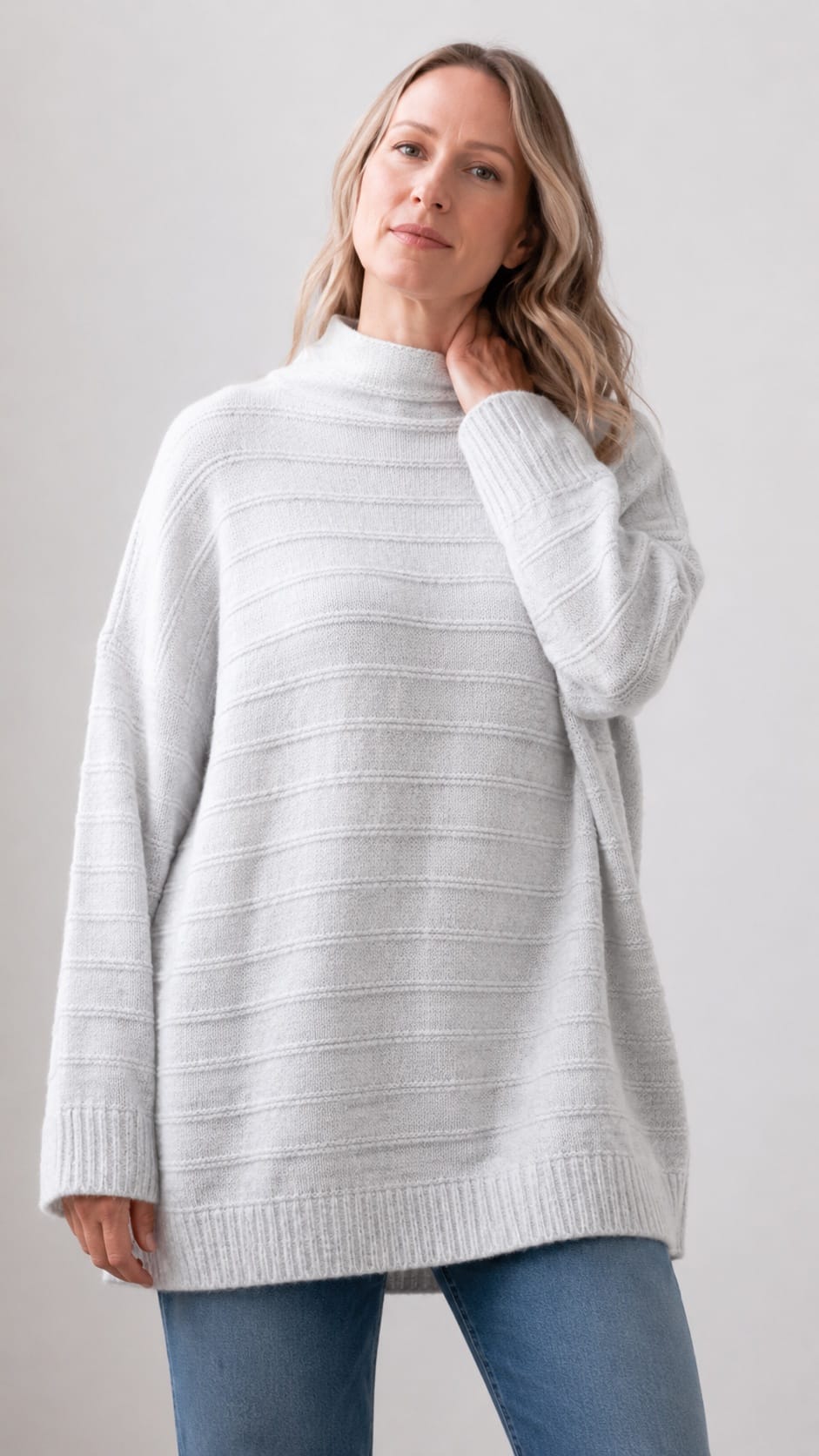 A woman wearing an oversized ribbed knit mock neck sweater in a light heather gray color, paired with blue jeans, against a plain light gray studio background.