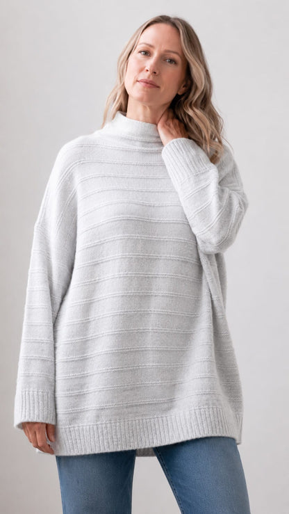A woman wearing an oversized ribbed knit mock neck sweater in a light heather gray color, paired with blue jeans, against a plain light gray studio background.