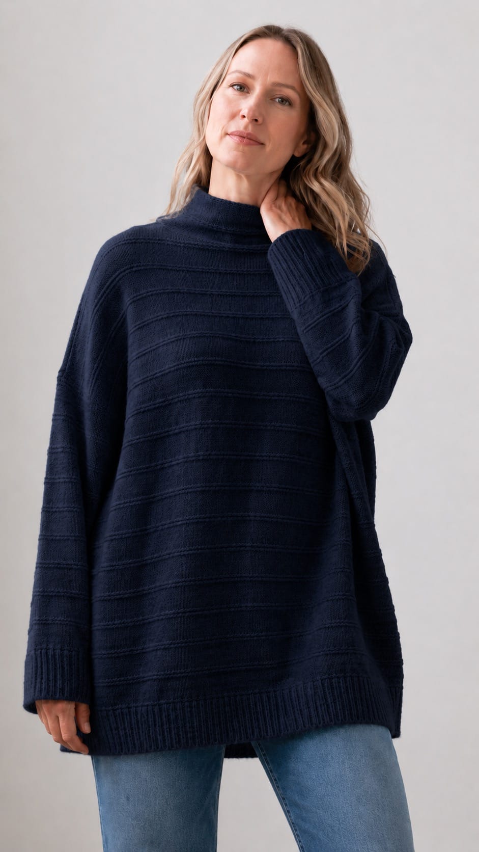 A woman wearing an oversized dark navy ribbed knit mock-neck sweater and light blue jeans, standing against a plain light gray studio background.