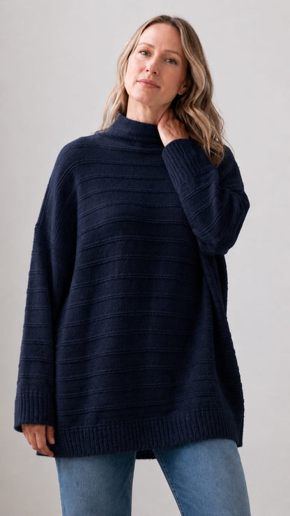 A woman wearing an oversized dark navy ribbed knit mock-neck sweater and light blue jeans, standing against a plain light gray studio background.