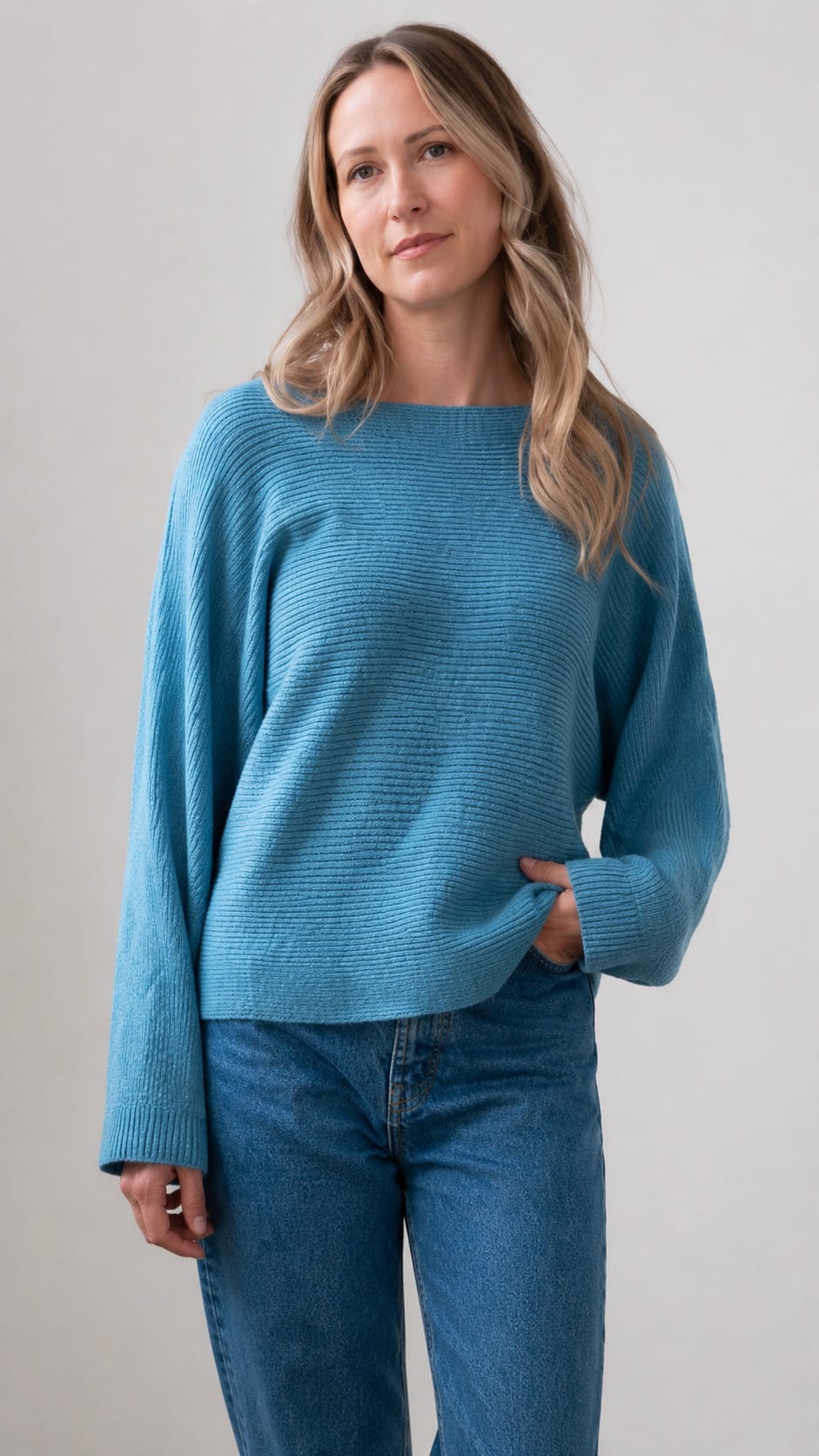 A woman wearing a textured knit long-sleeve sweater in a medium teal-blue color, paired with blue jeans, standing against a neutral light gray background.