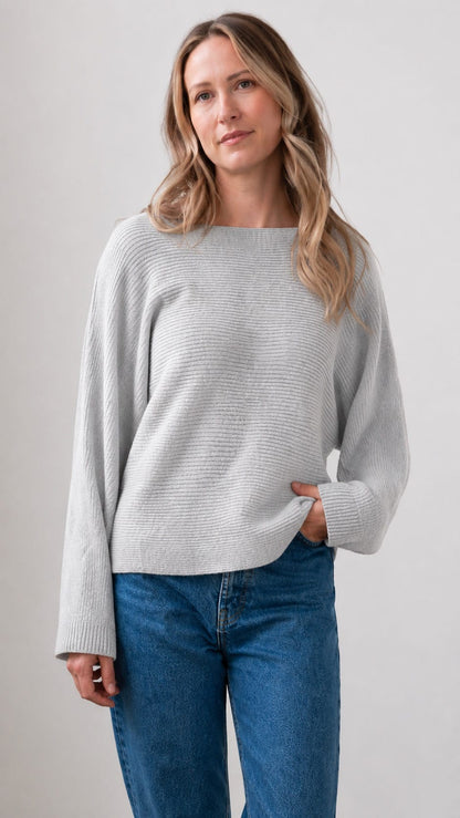 A woman wearing a light gray ribbed knit sweater with a boat neckline and blue jeans, standing against a plain light gray studio background.