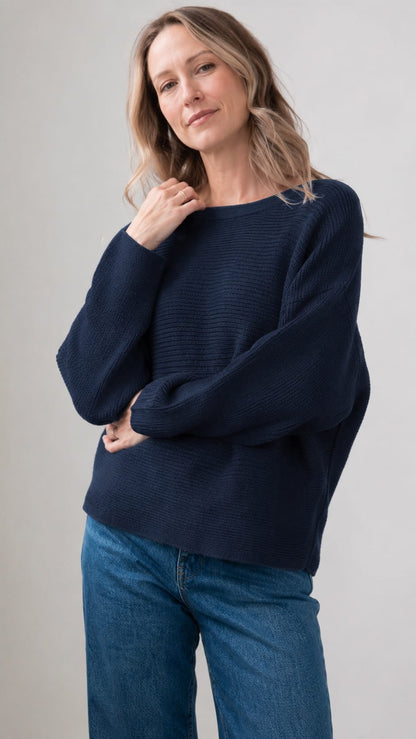 A woman wearing a dark navy blue ribbed knit sweater with balloon sleeves and blue jeans, standing against a neutral light gray background.