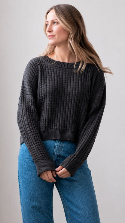 A woman wearing a dark charcoal gray ribbed knit sweater and blue jeans, standing against a light gray studio background.