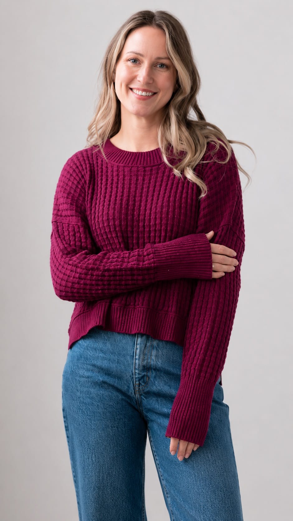 A woman wearing a dark magenta ribbed knit sweater and blue jeans, standing against a neutral light gray background.