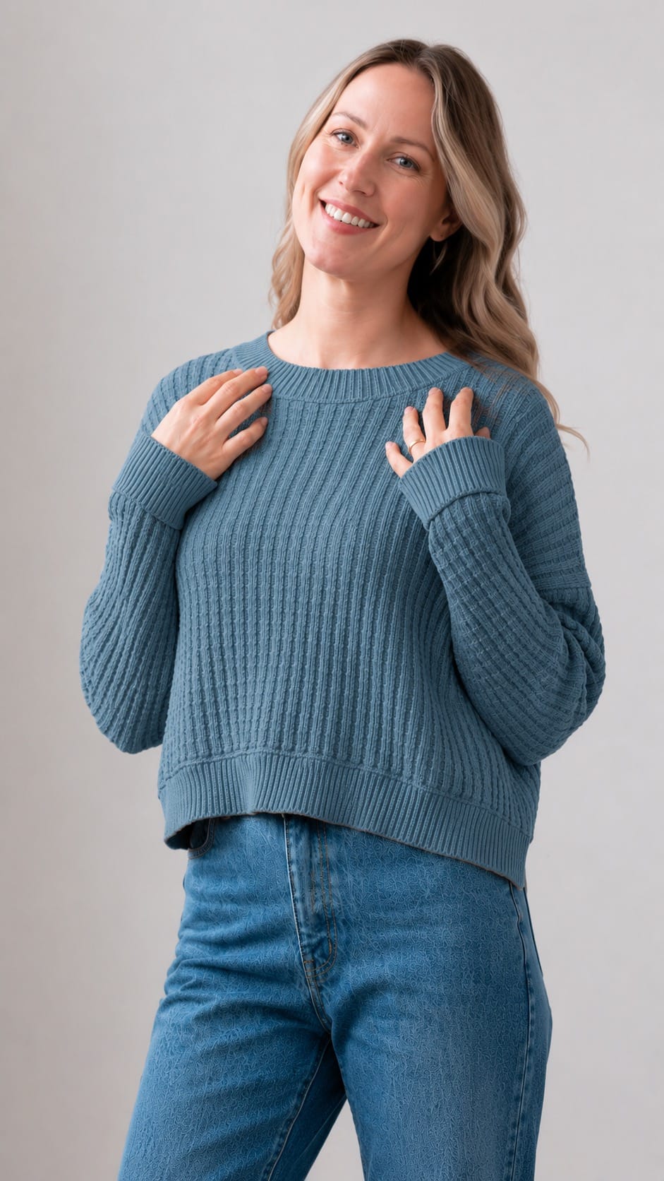  A woman wearing a medium blue ribbed knit sweater and blue jeans, standing against a light gray studio background.