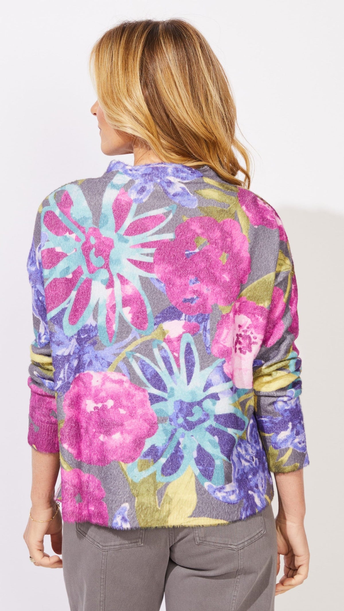 A woman shown from the back wearing a colorful floral print cardigan with pink, purple, and blue flowers on a gray background, paired with gray pants.
