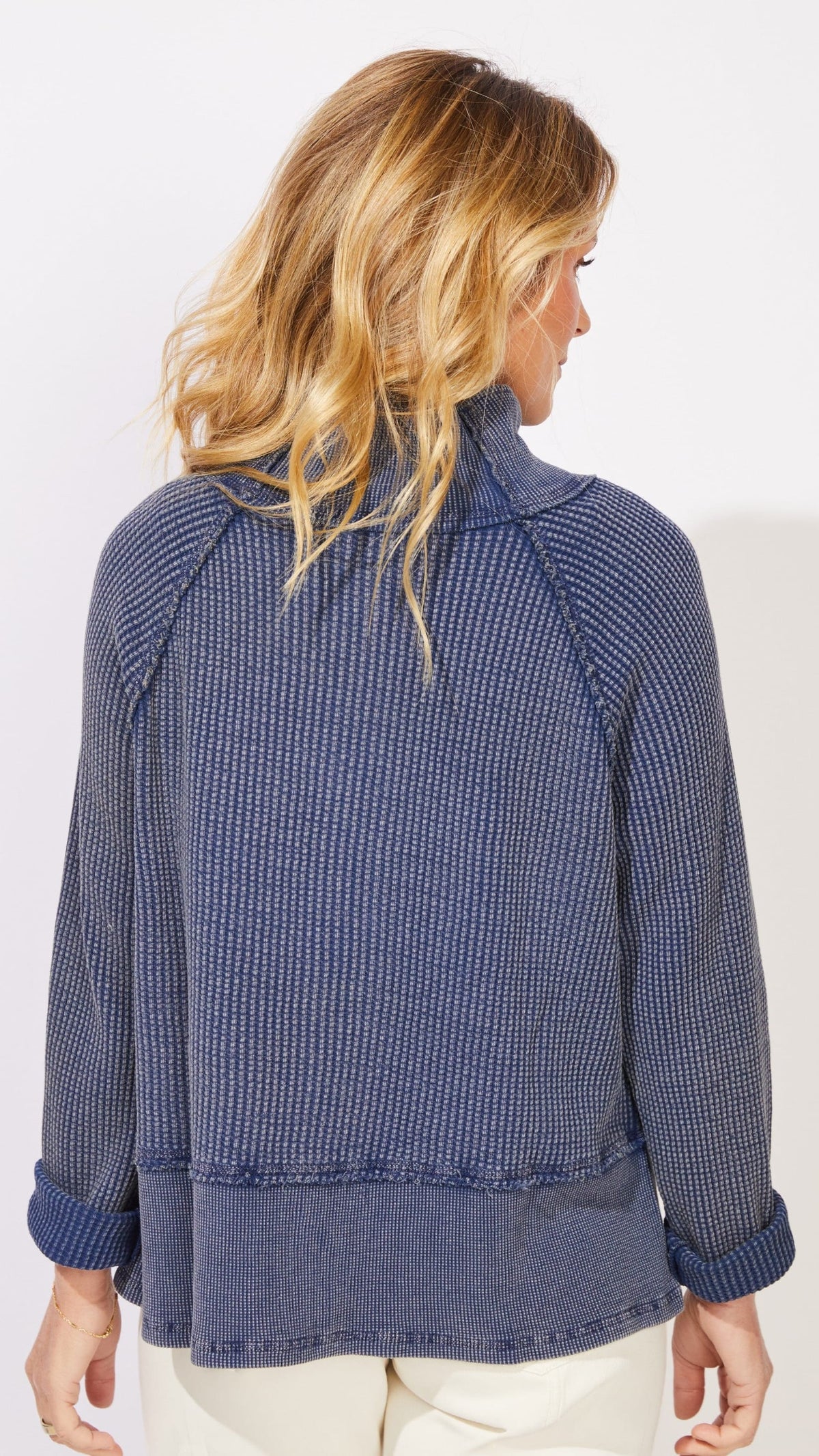 A woman shown from the back wearing a textured, blue-and-white vertically patterned long-sleeve top with rolled cuffs, paired with cream pants, against a plain white studio background.