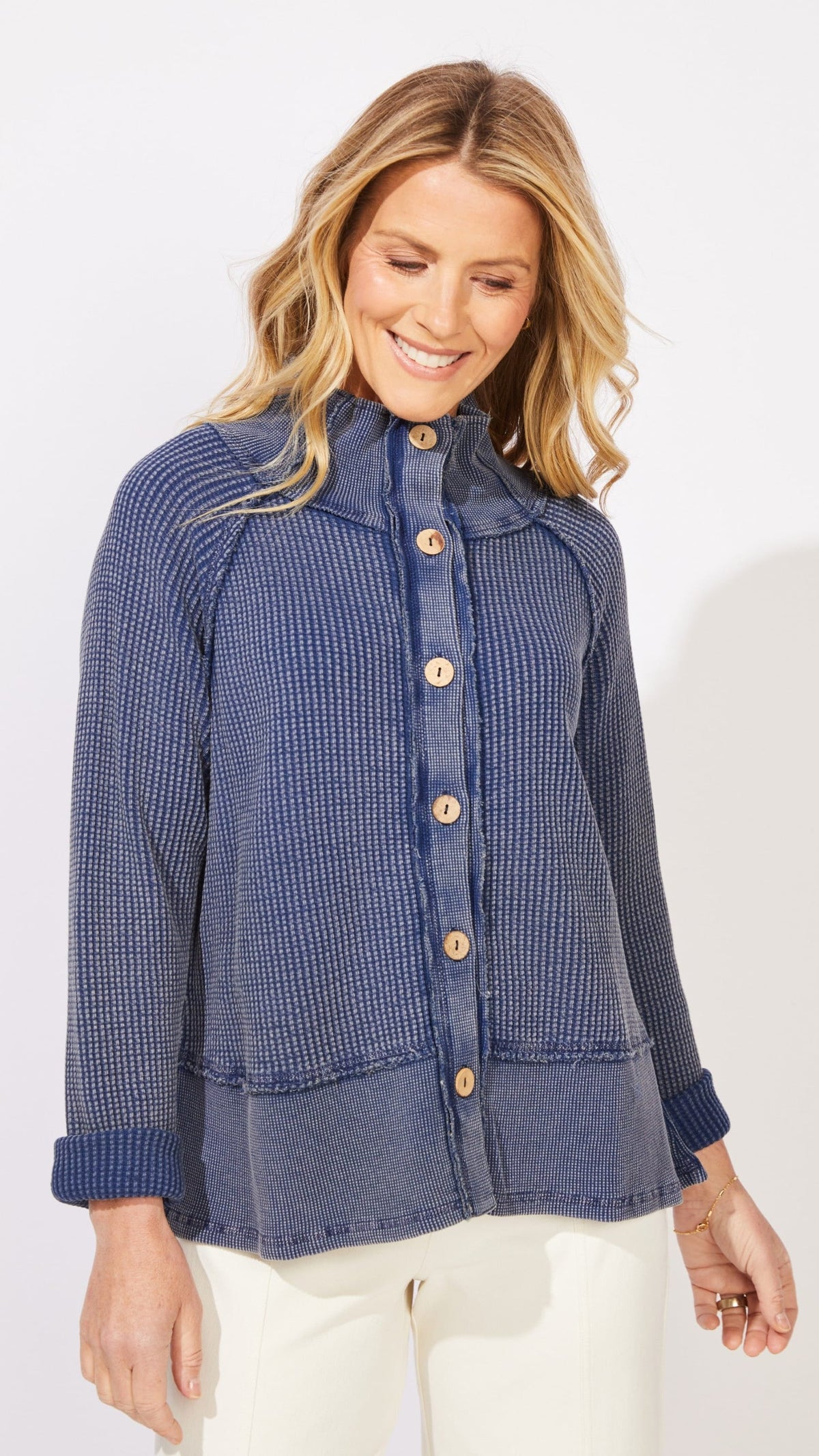 A woman wearing a blue ribbed button-front cardigan with a high collar and white pants, photographed against a clean white studio background.