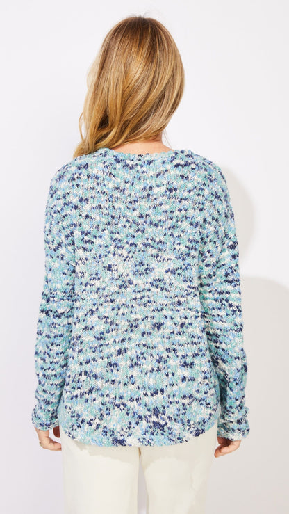 A woman viewed from the back wearing a long-sleeve textured sweater with a blue, navy, and white pattern, paired with cream pants, against a plain white background.