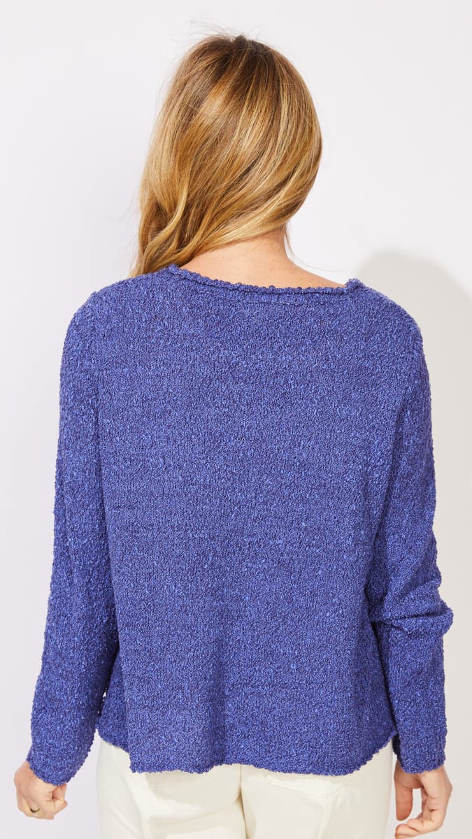 A woman viewed from behind wearing a textured, heathered blue-purple knit sweater and cream pants against a plain white background.