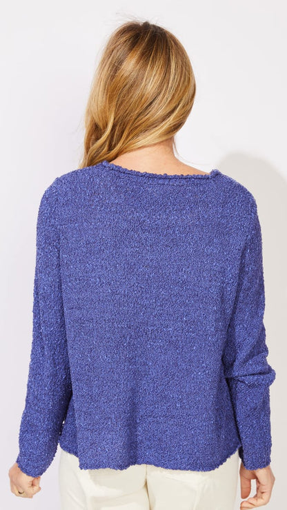 A woman viewed from behind wearing a textured, heathered blue-purple knit sweater and cream pants against a plain white background.