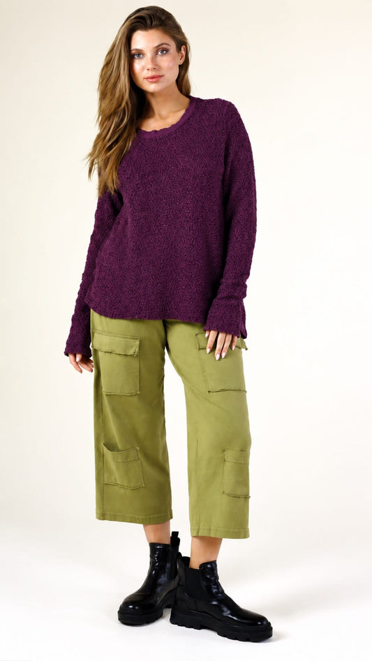 A woman modeling a fuzzy purple long-sleeve knit sweater paired with olive green cargo pants and black boots, standing against a plain off-white studio background.