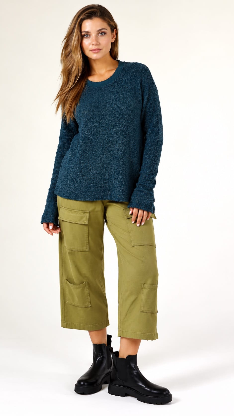 A woman modeling a dark teal textured knit sweater, olive green cropped cargo pants, and black Chelsea boots against a plain white studio background.