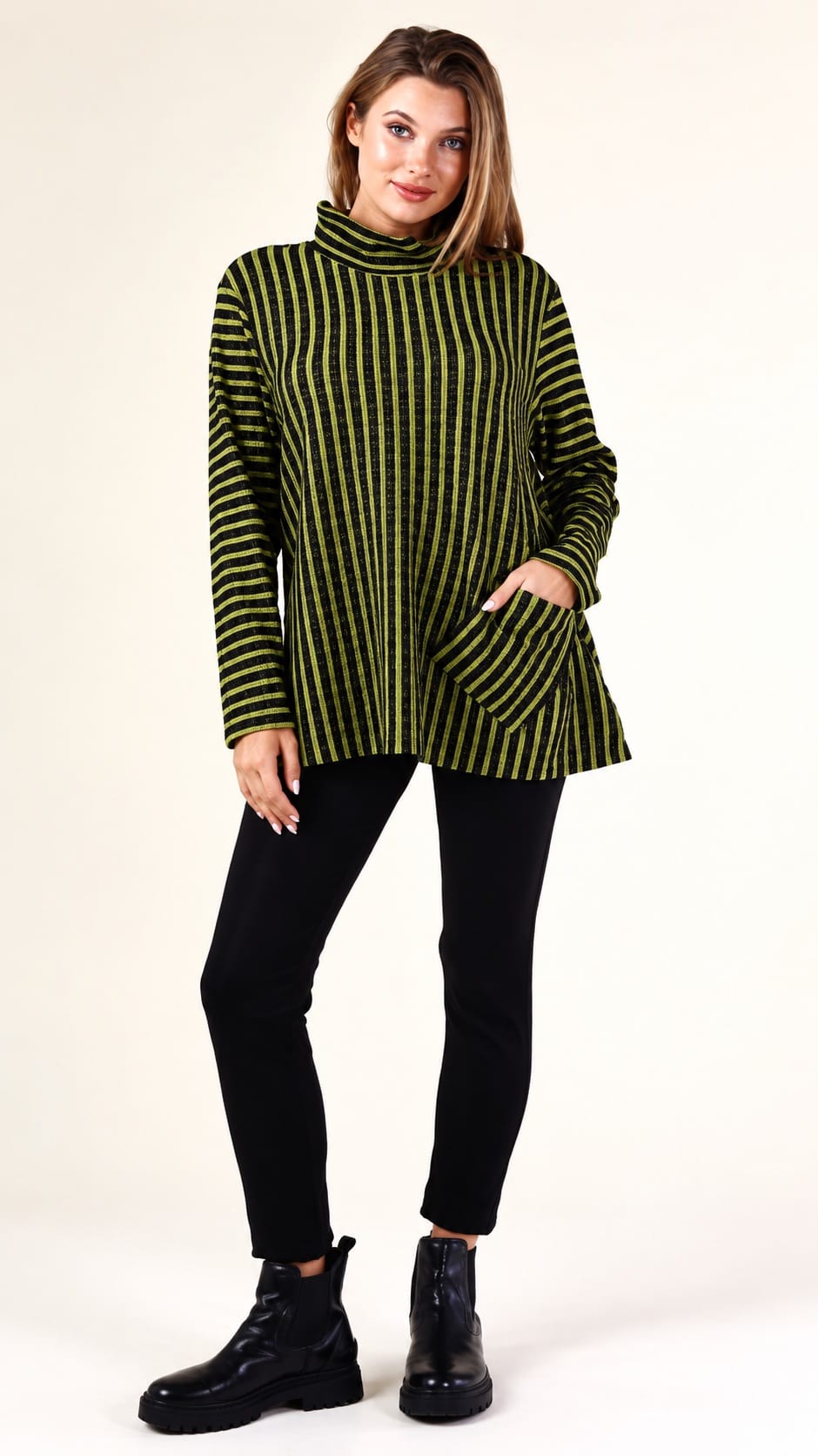 A woman modeling a green and black vertically striped turtleneck sweater with front pockets, paired with black pants and black boots, against a plain off-white studio background.