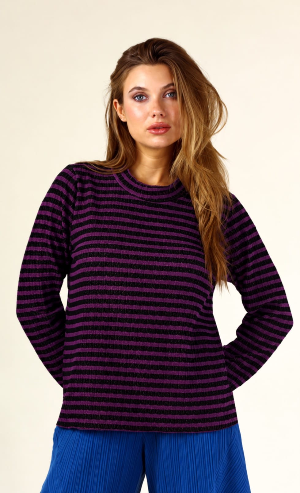 A woman modeling a dark purple and black horizontally striped long-sleeve knit top with blue pleated pants, set against a plain beige studio background.