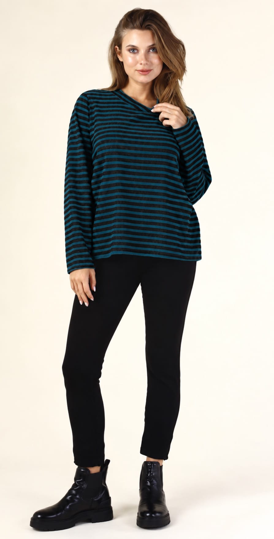A woman modeling a dark teal and black horizontally striped long-sleeve knit top, paired with black pants and black boots, against a plain cream studio background.