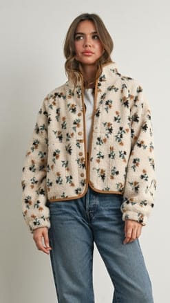 A woman modeling a cream-colored fuzzy teddy jacket with a brown and green floral pattern, paired with blue jeans, against a plain light gray background.