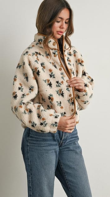 A woman wearing a cream-colored sherpa jacket with a brown and black floral pattern, paired with blue jeans, against a plain light gray background.