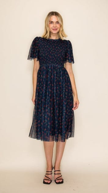 A woman modeling a dark navy blue midi dress with a small red and pink floral pattern, featuring short flutter sleeves and a smocked bodice, set against a plain beige background.