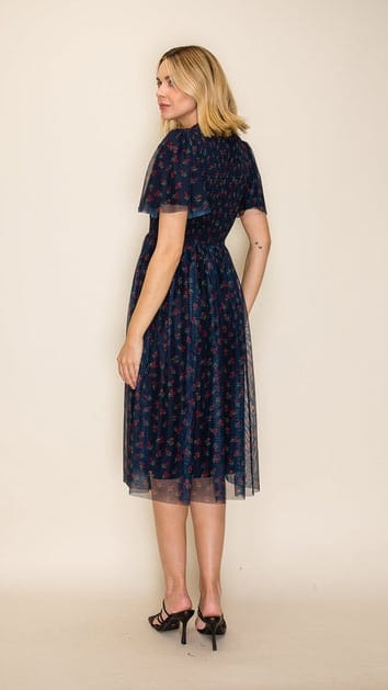 A woman modeling a dark navy midi dress with a red and white floral pattern, featuring short flutter sleeves and a sheer tulle overlay, shown from a back-three-quarter view against a plain beige studio background.