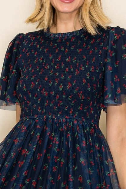 A close-up of a woman wearing a navy blue smocked dress with a red and green floral pattern, featuring sheer flutter sleeves.