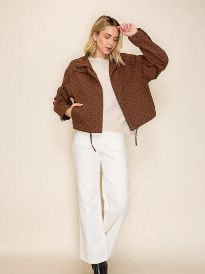 A woman modeling a cropped brown jacket with a subtle black polka dot pattern, worn over a cream top and white wide-leg pants, set against a plain beige studio background.