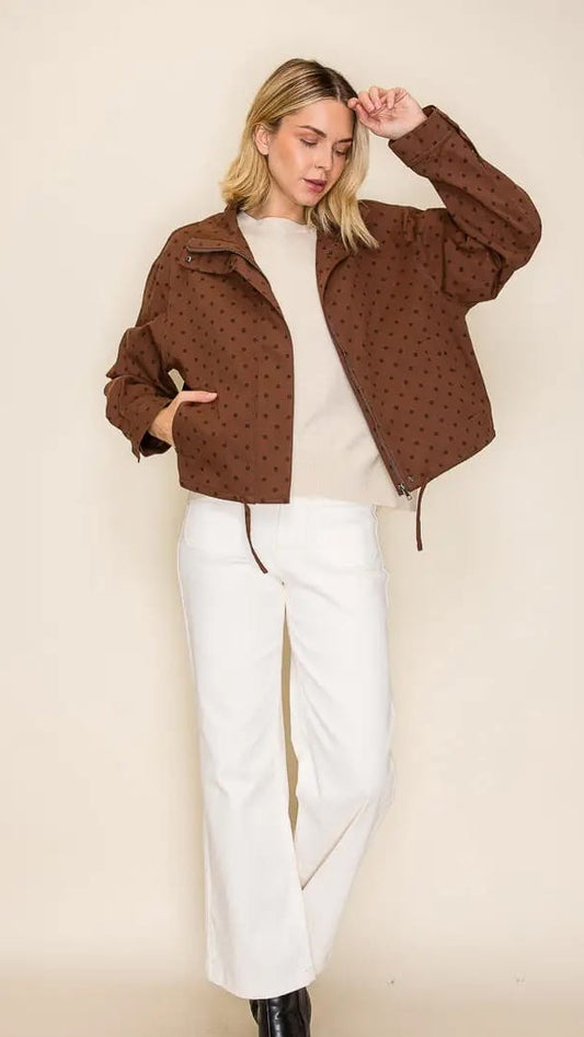 A woman modeling a cropped brown jacket with a subtle black polka dot pattern, worn over a cream top and white wide-leg pants, set against a plain beige studio background.