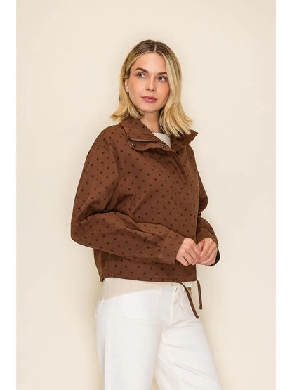 A woman modeling a brown long-sleeve cropped jacket with a small repeating polka dot pattern, paired with white pants, against a solid beige studio background.