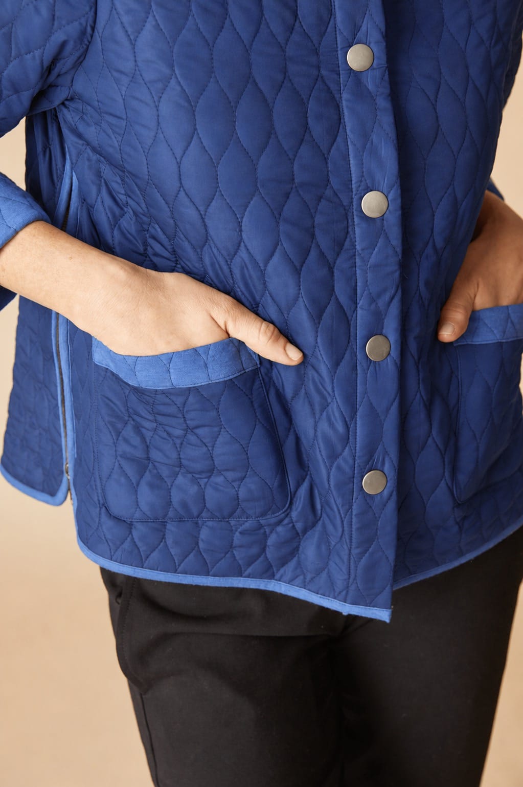 A close-up of a person wearing a quilted royal blue jacket with silver buttons and black pants, with their hands tucked into the jacket's front pockets.