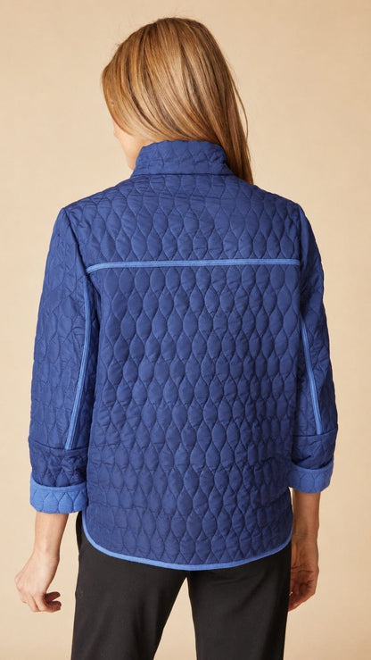 A woman viewed from the back wearing a blue quilted jacket with a diamond pattern and light blue piping details, paired with black pants, against a solid beige background.