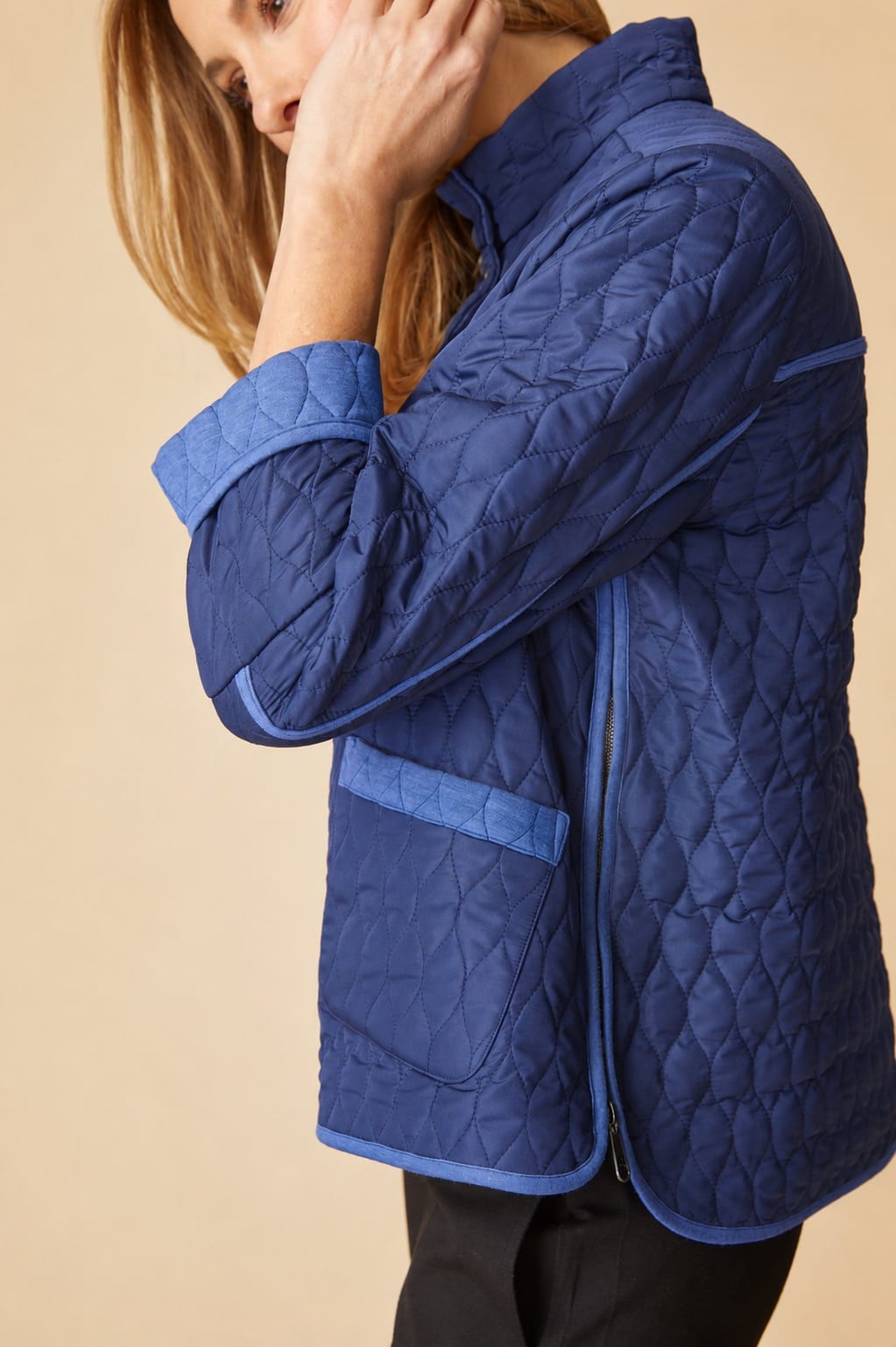 A woman wearing a blue quilted jacket with matching blue trim, shown from the side against a plain beige background.