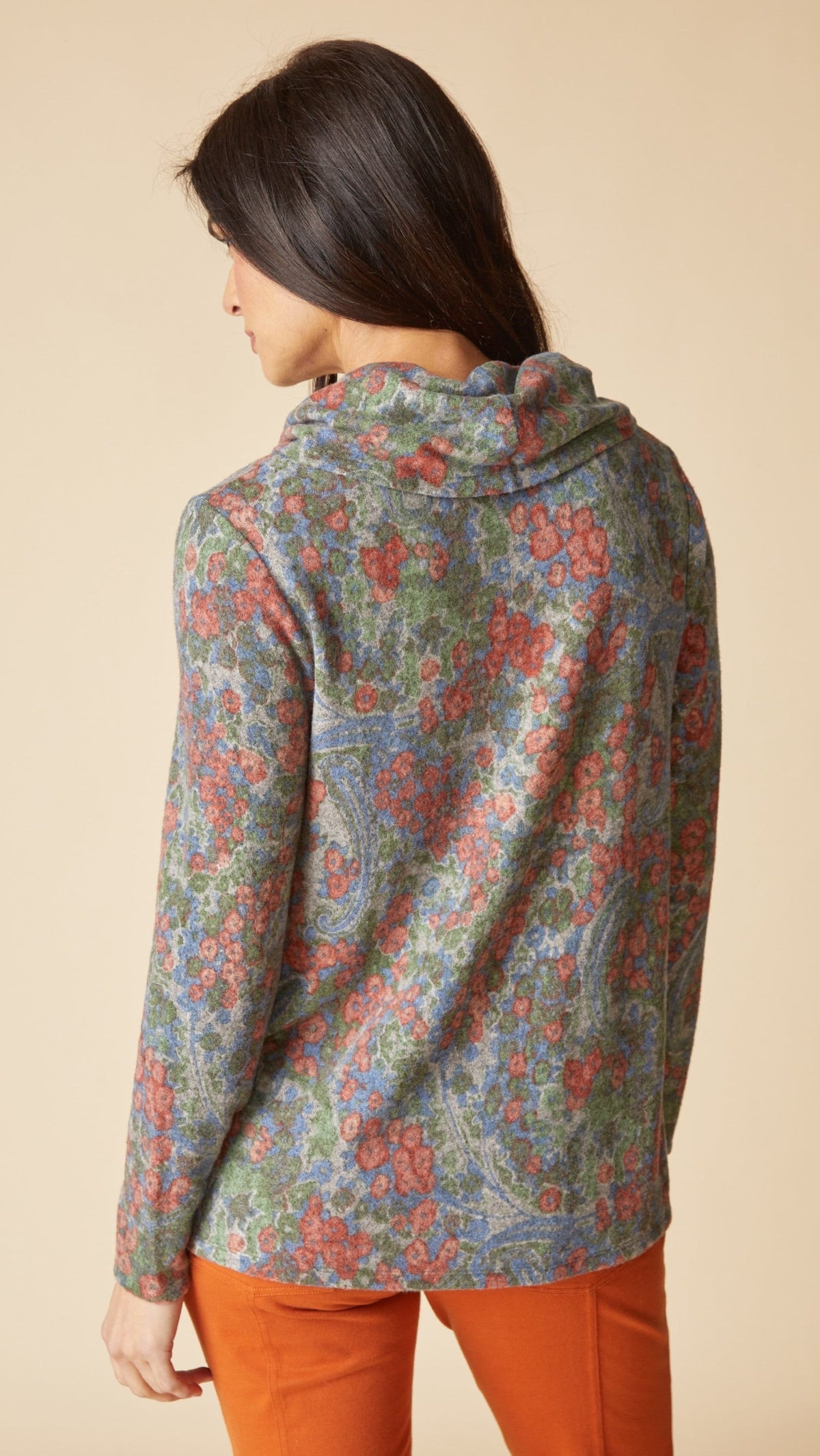 A woman shown from the back wearing a long-sleeve hooded top with an intricate multicolor paisley pattern in muted blue, green, and red tones, paired with orange pants, against a solid beige background.