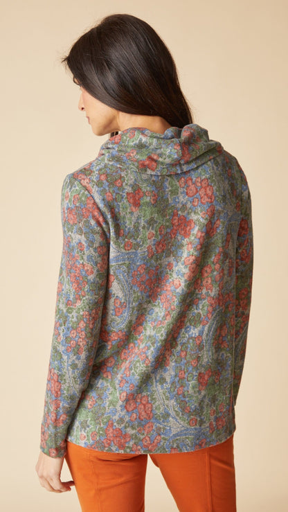 A woman shown from the back wearing a long-sleeve hooded top with an intricate multicolor paisley pattern in muted blue, green, and red tones, paired with orange pants, against a solid beige background.