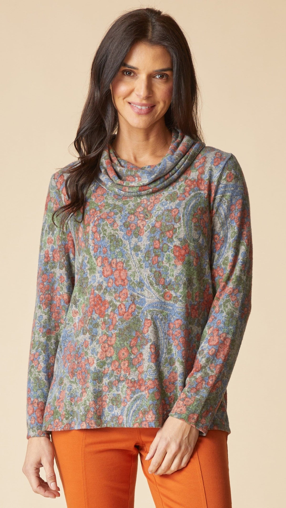 A woman modeling a long-sleeve cowl neck top with a multicolor paisley and floral pattern in muted blue, green, and coral tones, paired with orange pants, against a solid beige background.