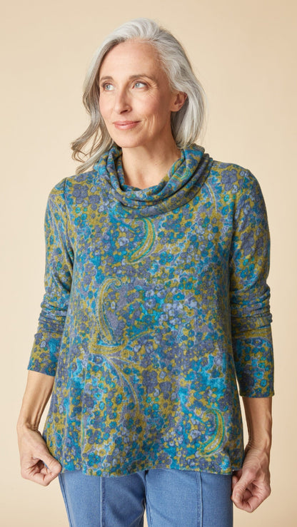 A woman with gray hair wearing a long-sleeve cowl neck top with a blue, green, and yellow paisley pattern, paired with blue jeans, against a solid beige background.