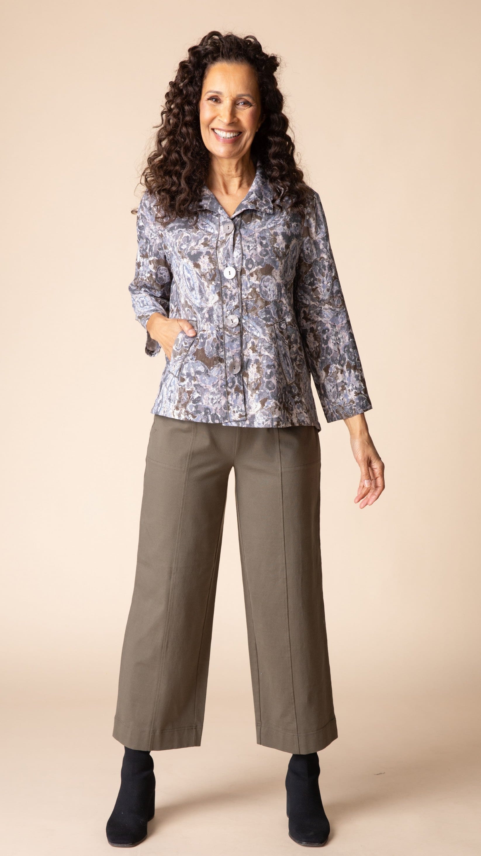 A smiling woman with curly dark hair stands against a plain beige studio background wearing a patterned button-front blouse with long sleeves and olive wide-leg cropped pants with black ankle boots.