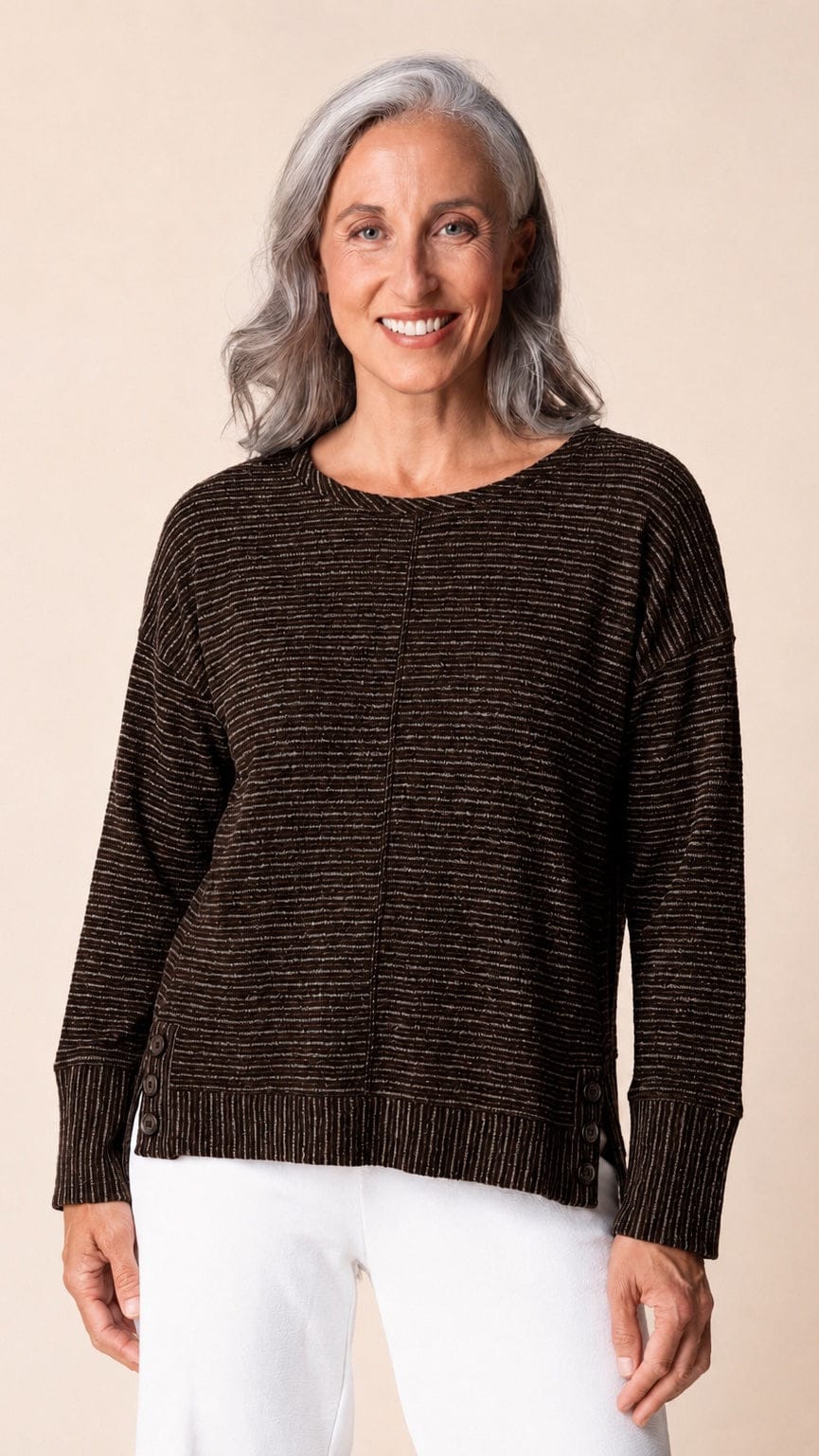 A smiling woman with gray hair wearing a dark brown long-sleeve knit sweater with thin horizontal metallic gold stripes, paired with white pants, against a solid beige background.