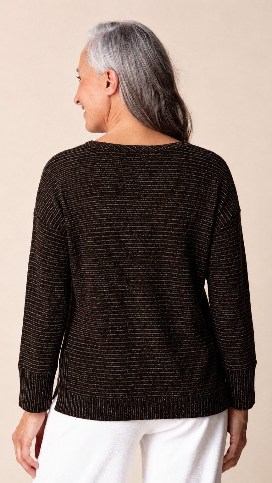An older woman with gray hair is shown from the back wearing a dark brown textured knit sweater with thin horizontal stripes and white pants, set against a plain beige background.