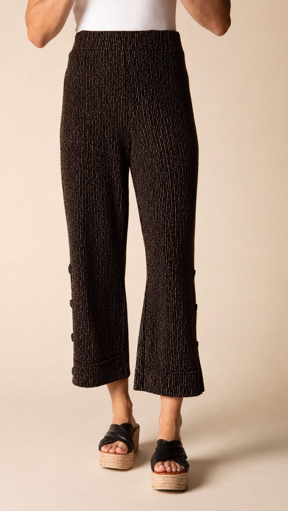 A person wearing textured black cropped pants with side buttons and black espadrille sandals, shown from the waist down against a plain beige studio background.