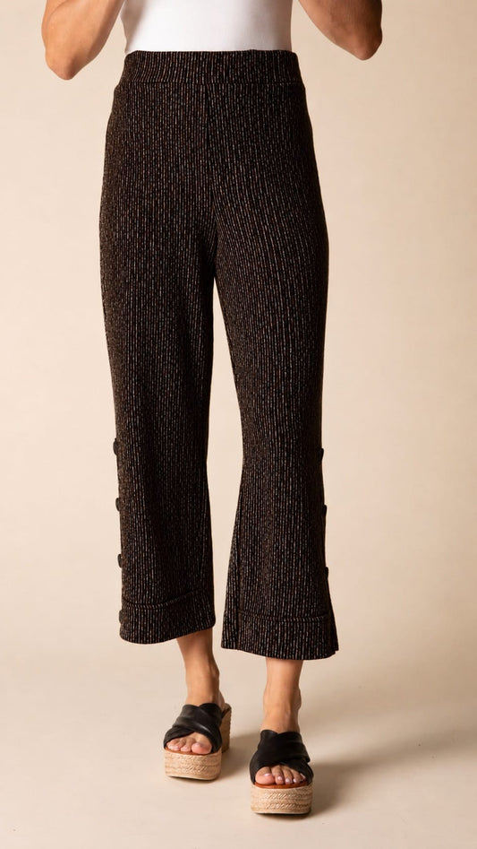 A person wearing textured black cropped pants with side buttons and black espadrille sandals, shown from the waist down against a plain beige studio background.