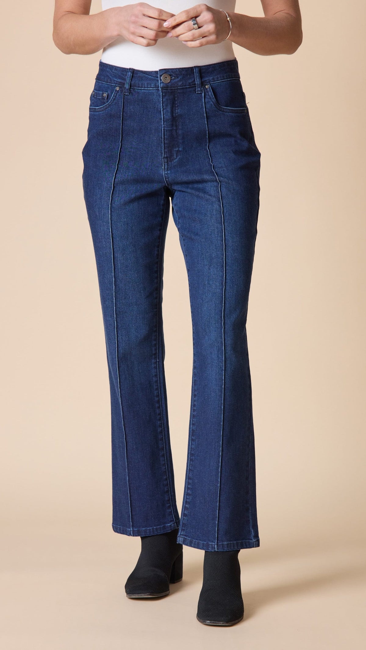 A person wearing dark blue high-rise flare jeans with front seam detailing, paired with a white top and black ankle boots, standing against a solid beige background.